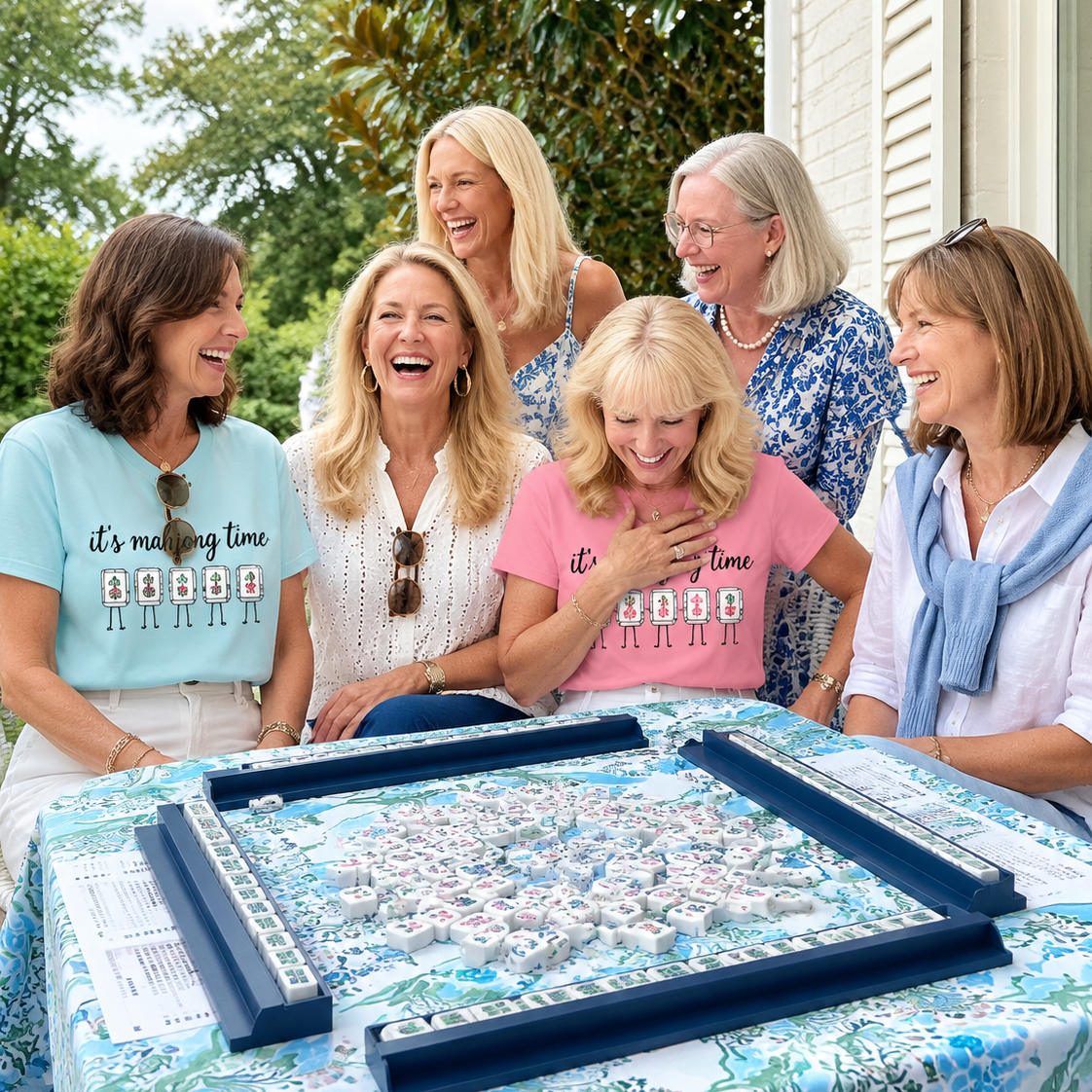 Five women laughing together outdoors behind a table covered with a blue and white floral tablecloth. On the table is a large rectangular game board with blue frames and many small white pieces.