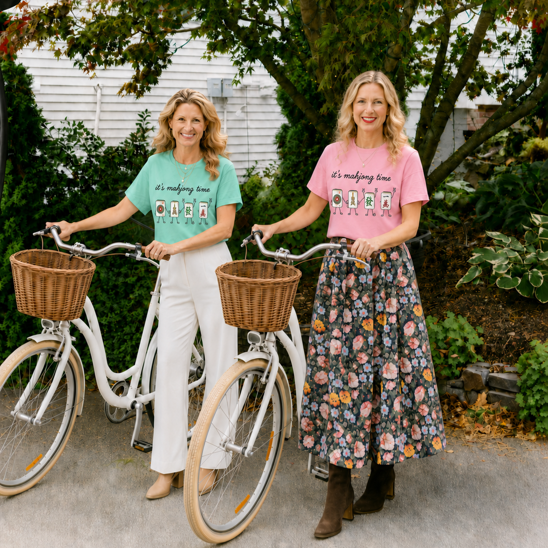 Two women standing outdoors with bicycles, wearing matching graphic t-shirts. The woman on the left wears a mint green shirt with 'it's making time OHO OHO' and white pants.