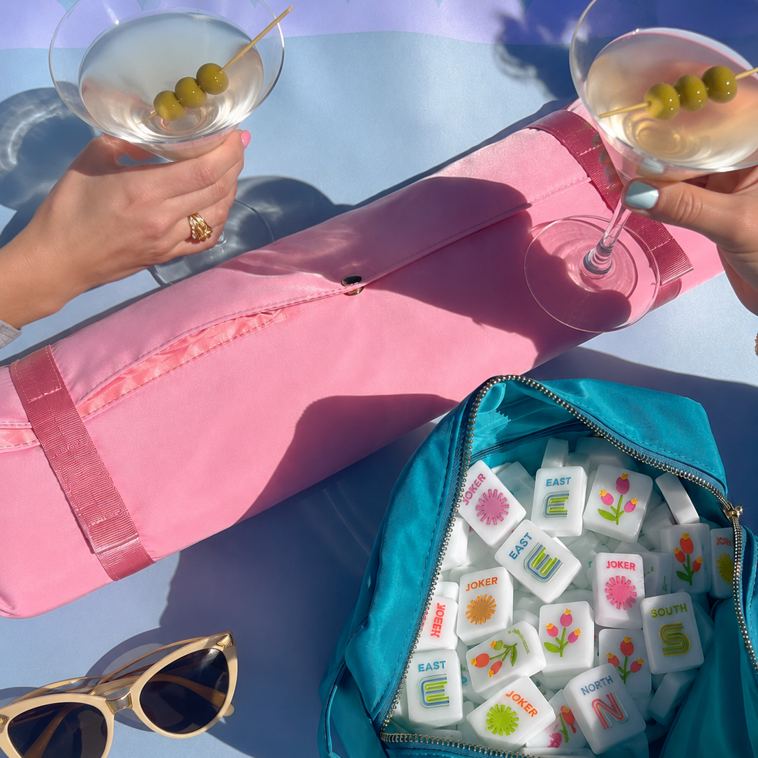 A pink rolled exercise/yoga mat and a teal zip pouch filled with white game pieces are arranged on a pastel lavender surface, with two hands holding cocktail glasses and sunglasses nearby.